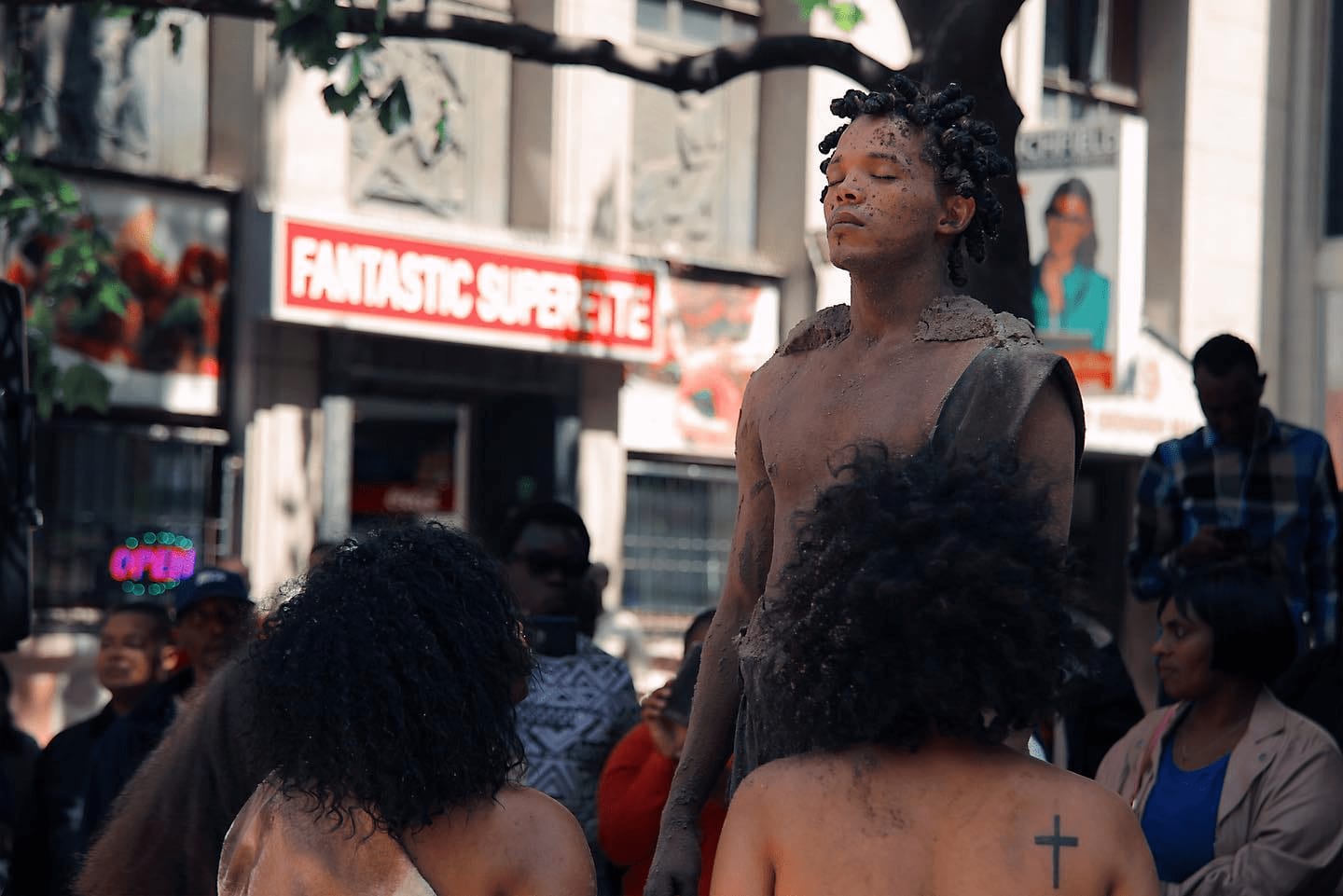 an image od an african mixed race man dressed in brown artistic rags and standing with his eyes closed performing an art with his group gathered around him and a crowd watching them